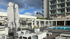 A rooftop pool area with lounge chairs and umbrellas, surrounded by a modern building with balconies.