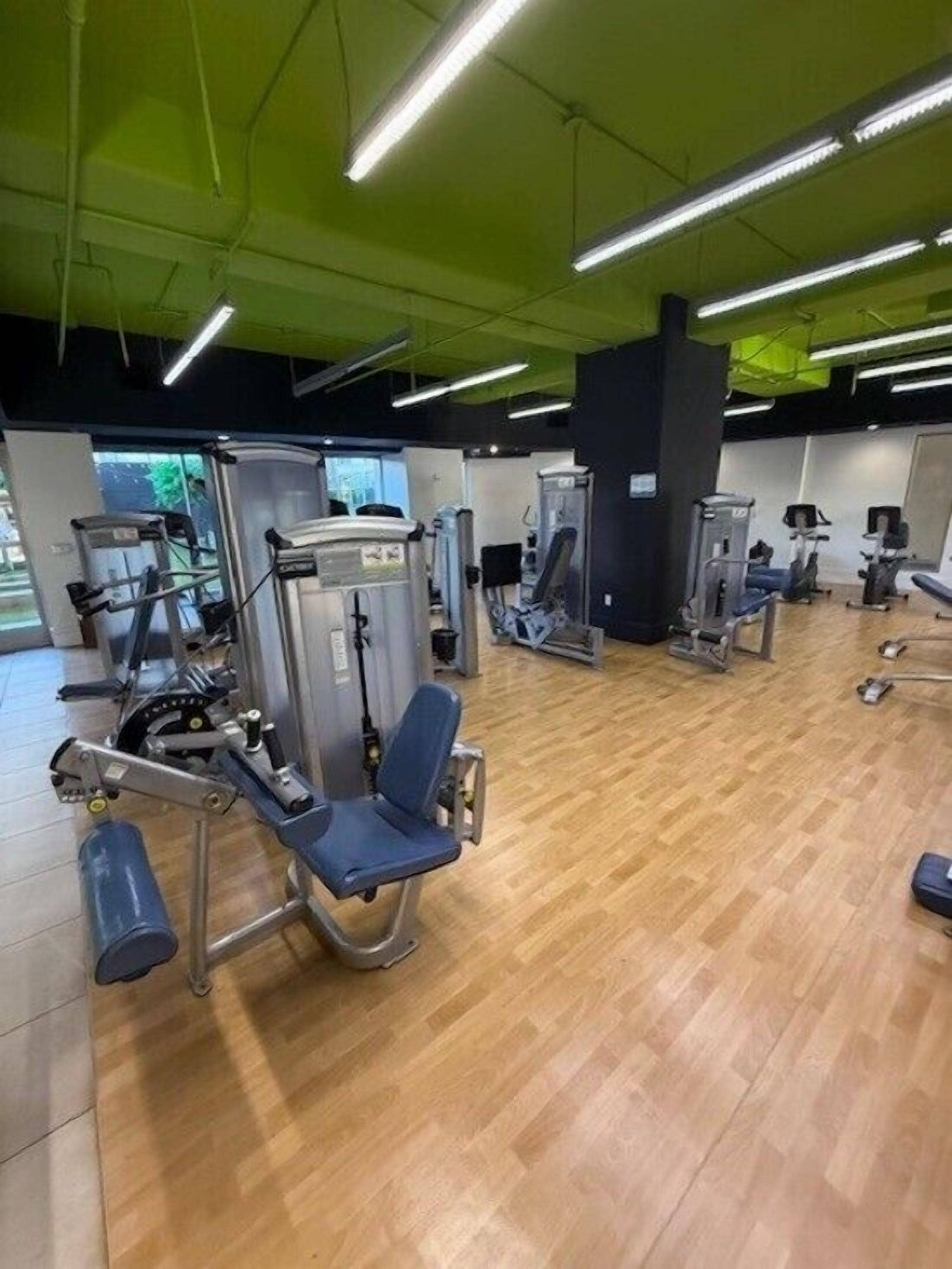 A gym with various exercise machines, wooden flooring, and green ceiling lights.