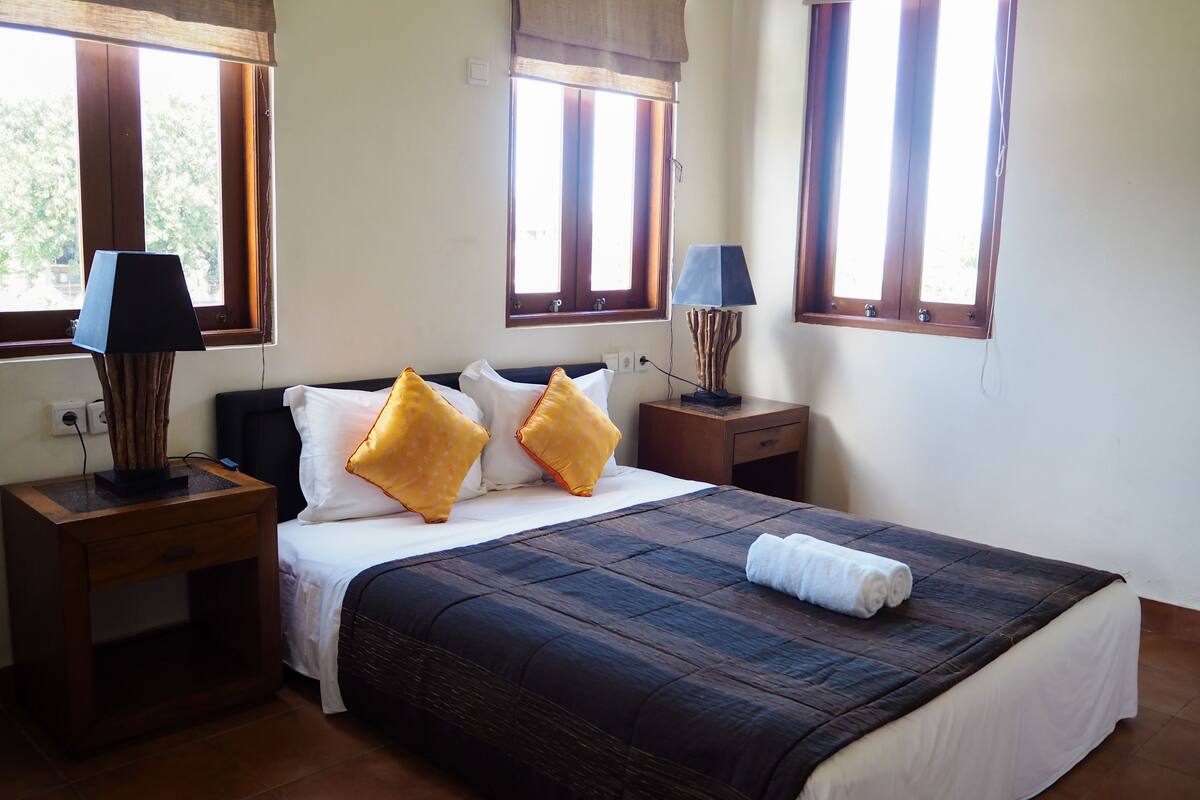 A neatly made bed with a dark quilt, two bedside lamps, and a window with a curtain.