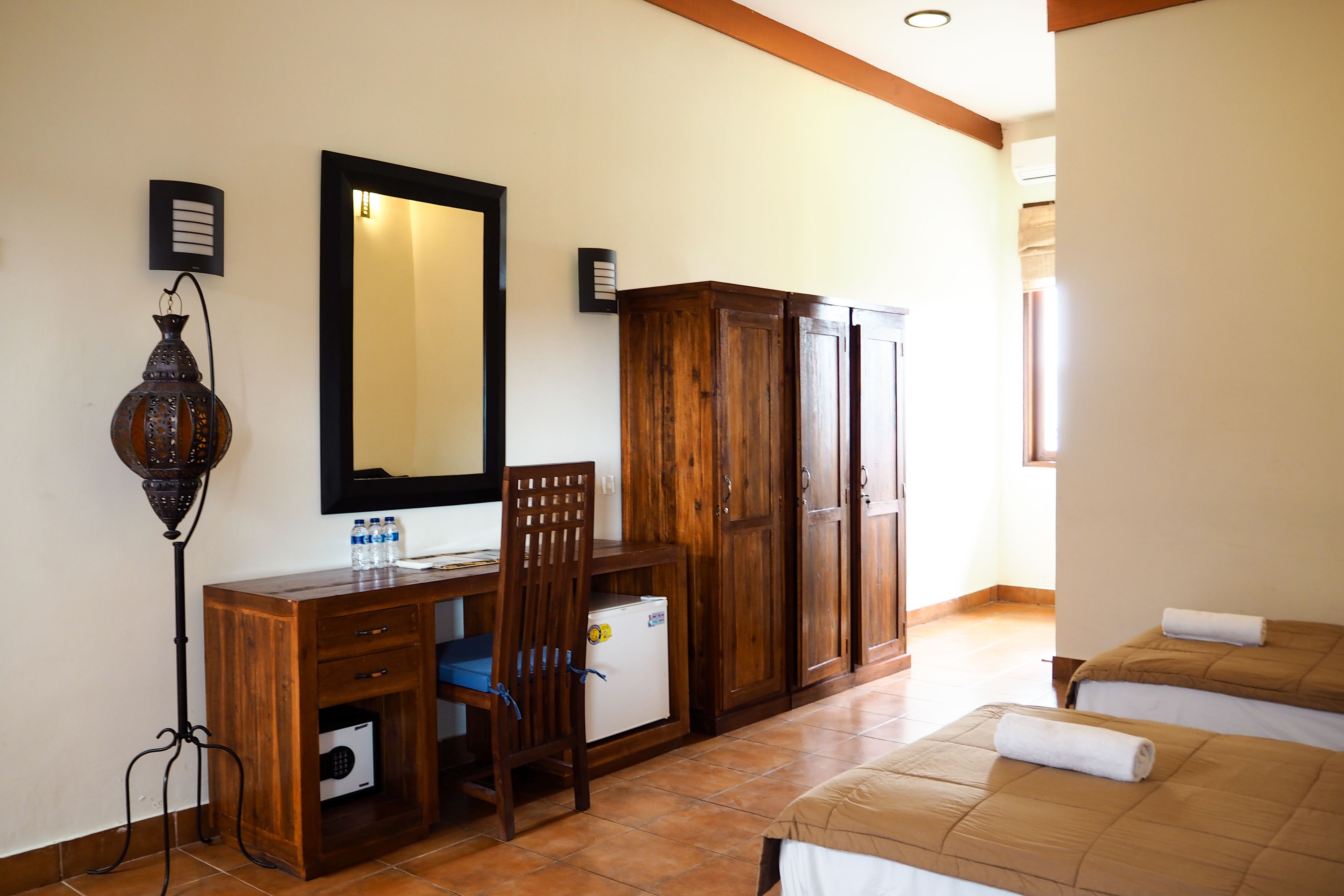 Une chambre d’hôtel avec un bureau, une chaise et une armoire en bois, un lit recouvert d’une courtepointe marron et un grand miroir au mur.