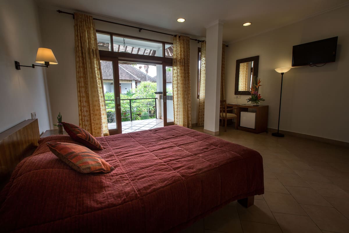 A bedroom with a large bed, a desk, a chair, a TV, and a balcony with curtains.