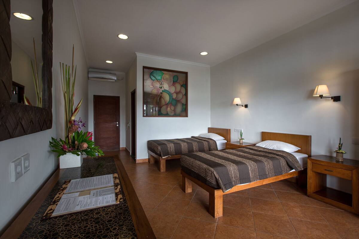 A hotel room with two beds, a wooden headboard, a nightstand, a small table with a plant, and a painting on the wall.
