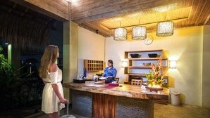 A reception area with a wooden reception desk, a clock, and a reception desk attendant. There is a plant and a vase with flowers on the desk. - Ecosfera Hotel (Canggu)