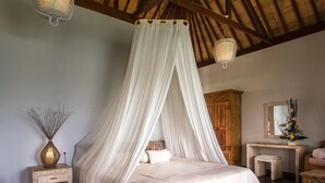 A bedroom with a four-poster bed, wooden flooring, a wooden wardrobe, a bedside table with a vase, and a mirror on the wall. - Ecosfera Hotel (Canggu)
