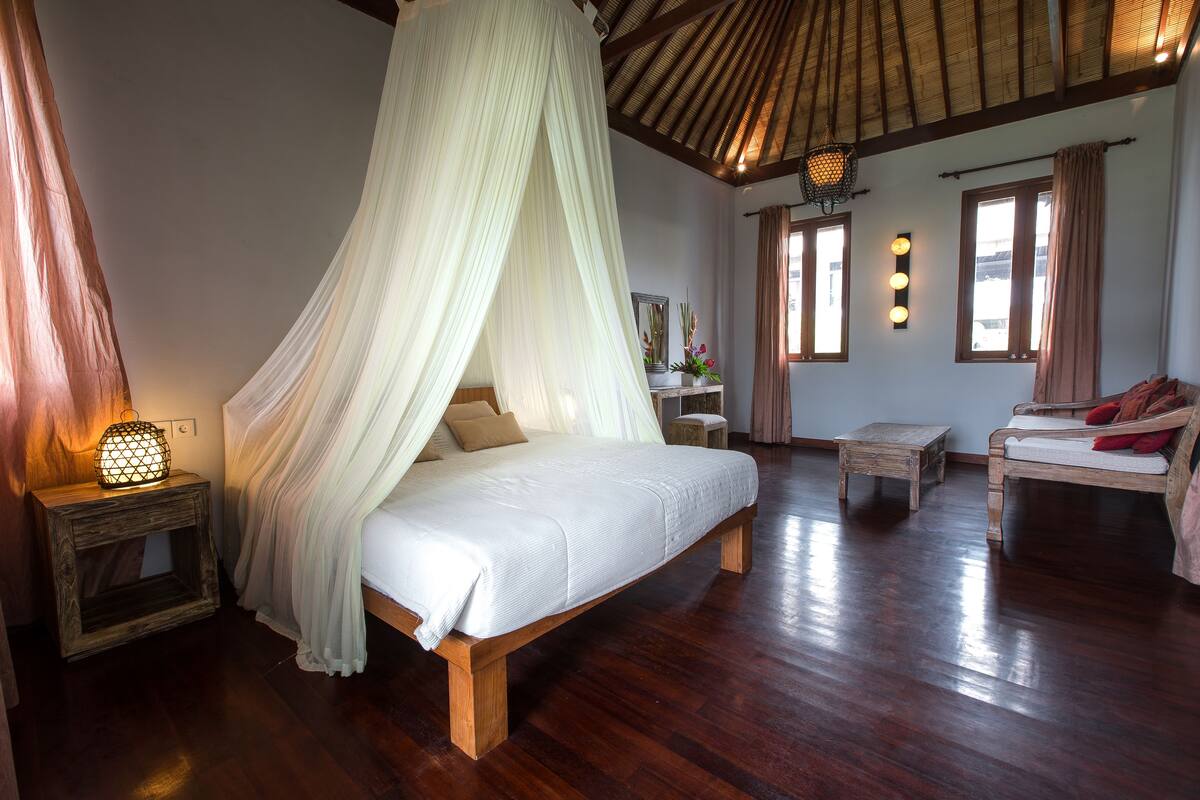 A bedroom with a canopy bed, wooden flooring, a nightstand with a lamp, a bedside table, and a sofa.
