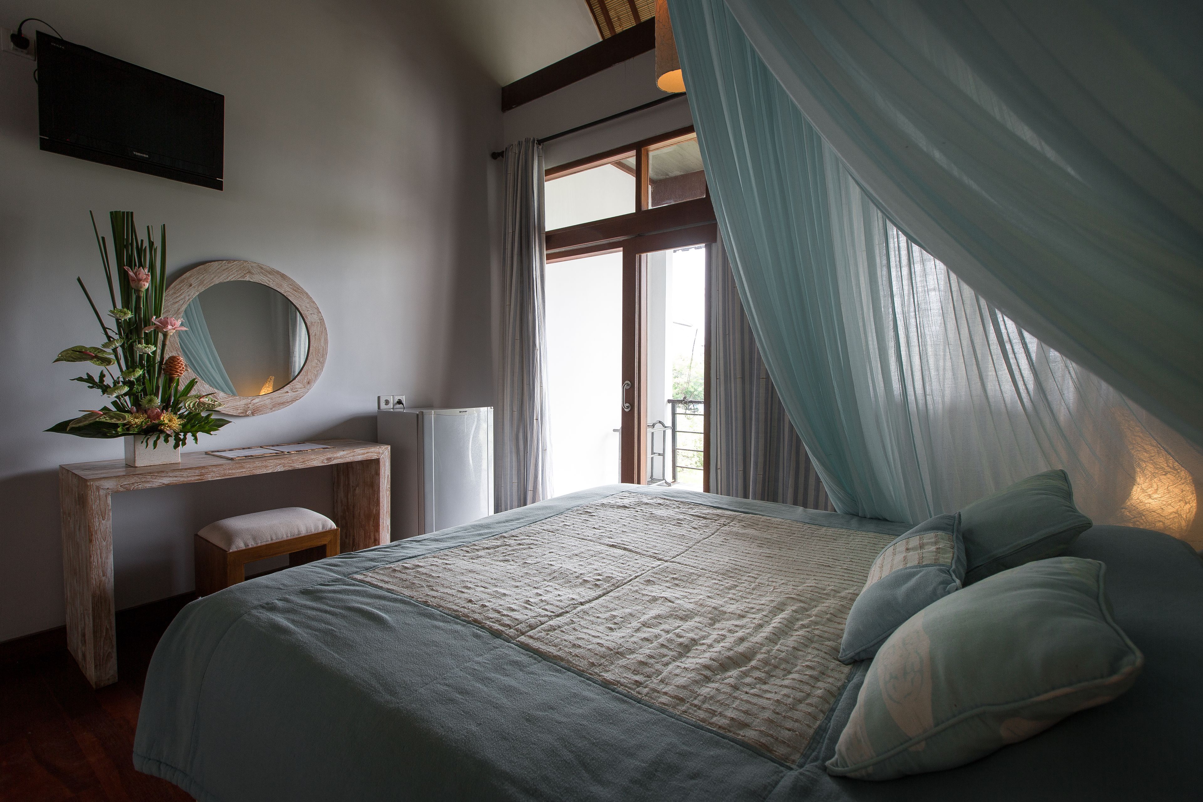 A bedroom with a bed, a wooden desk, a mirror, and a window with curtains.