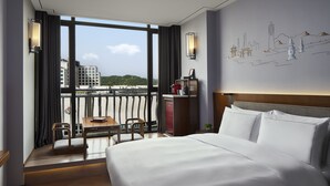 A hotel room with a large bed, a desk, a chair, and a view of a cityscape through large windows.