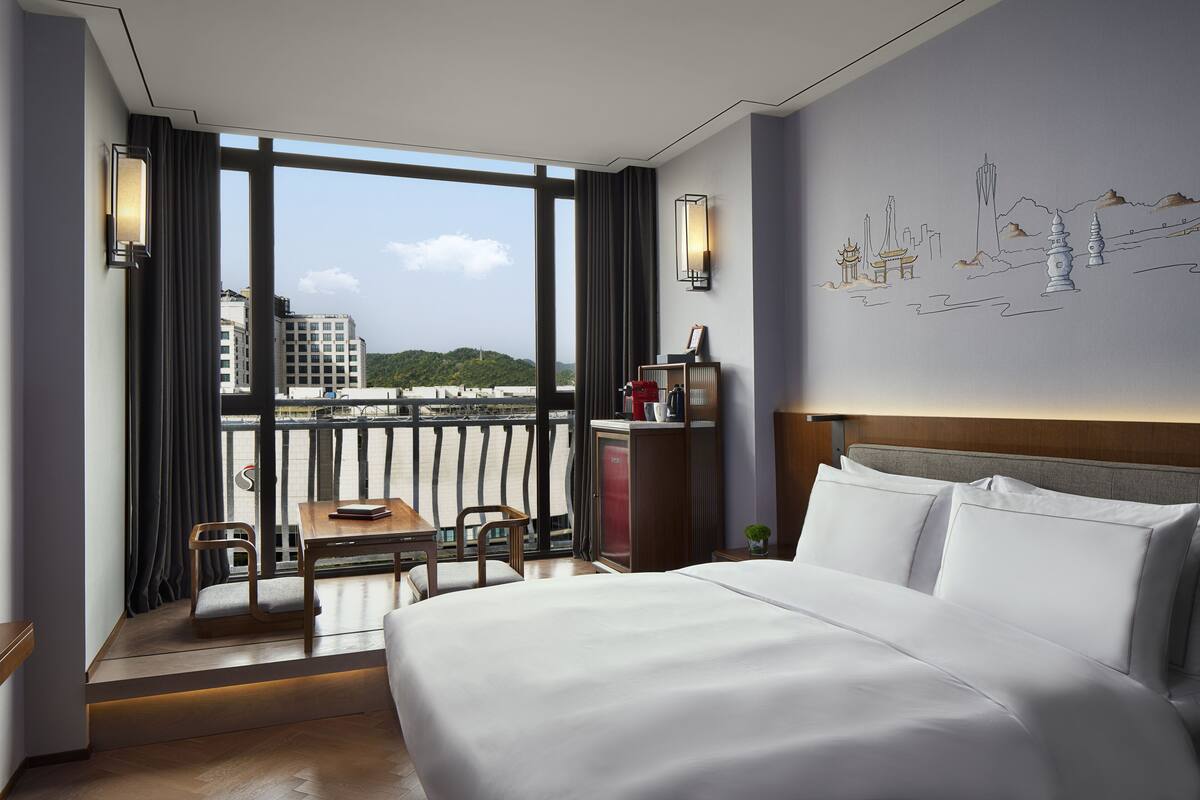 A hotel room with a large bed, a desk, a chair, and a view of a cityscape through large windows.