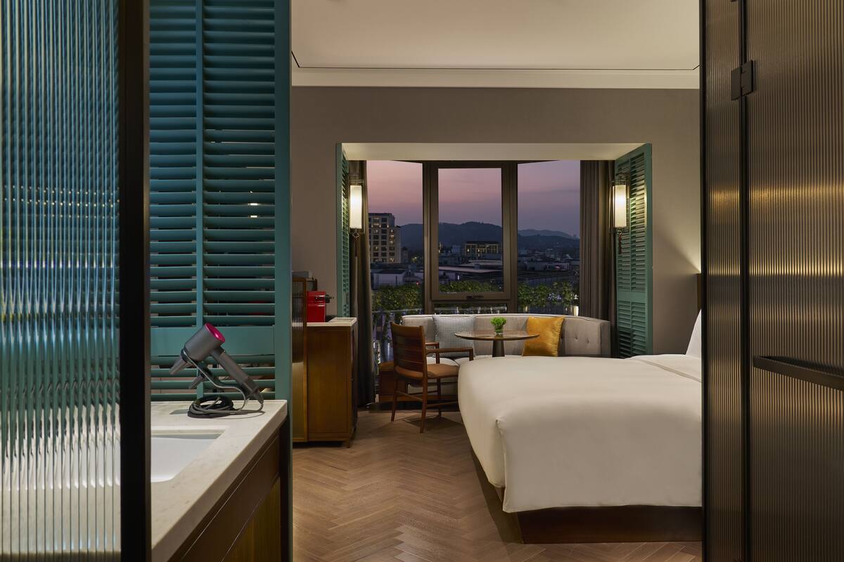 A hotel room with a bed, a desk with a lamp, a chair, and a view of the cityscape through large windows.