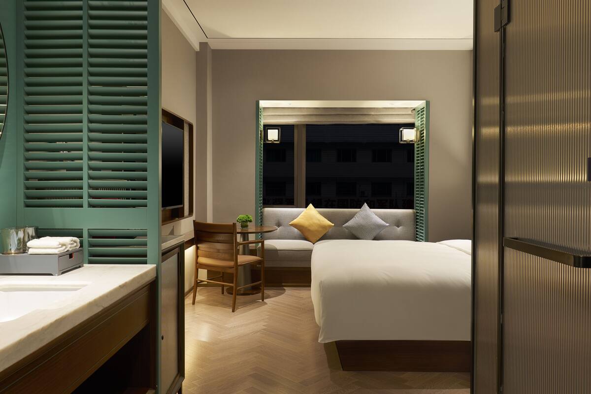 A hotel room with a bed, a desk with a chair, a window with a view of the outside, and a bathroom with a mirror and sink.