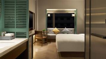 A hotel room with a bed, a desk with a chair, a window with a view of the outside, and a bathroom with a mirror and sink.