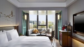 A hotel room with a bed, a seating area with a table, a TV, and a view of the city through large windows.