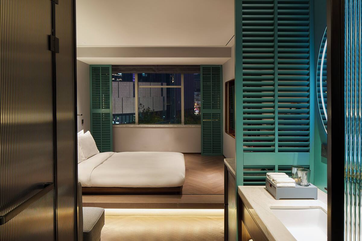 A modern bedroom with a large bed, a window with shutters, and a bedside table with a lamp.