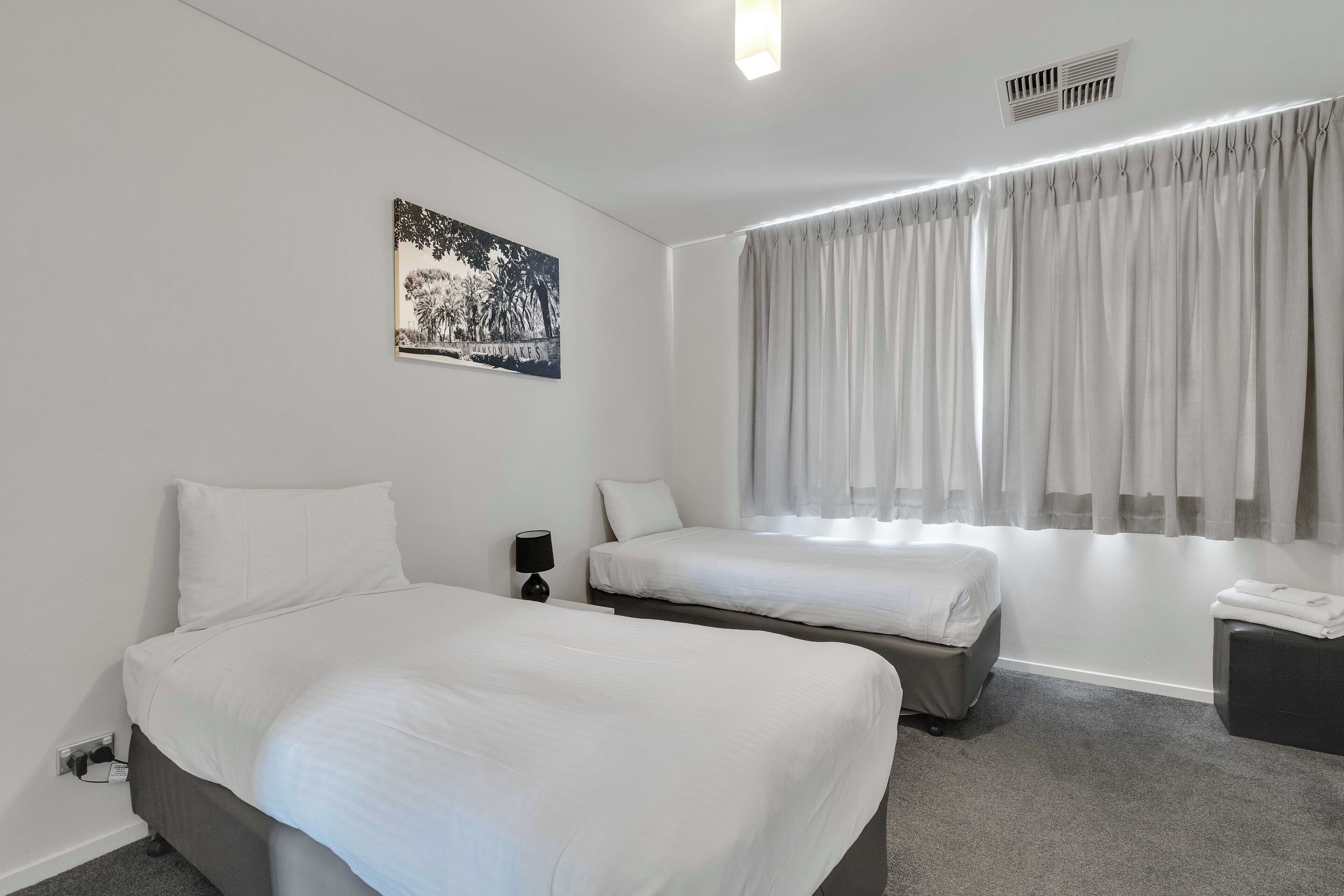 Hotellihuone, jossa on kaksi sänkyä, yöpöytä lampun kanssa, ikkuna verhoilla ja seinälle ripustettu taulu.
