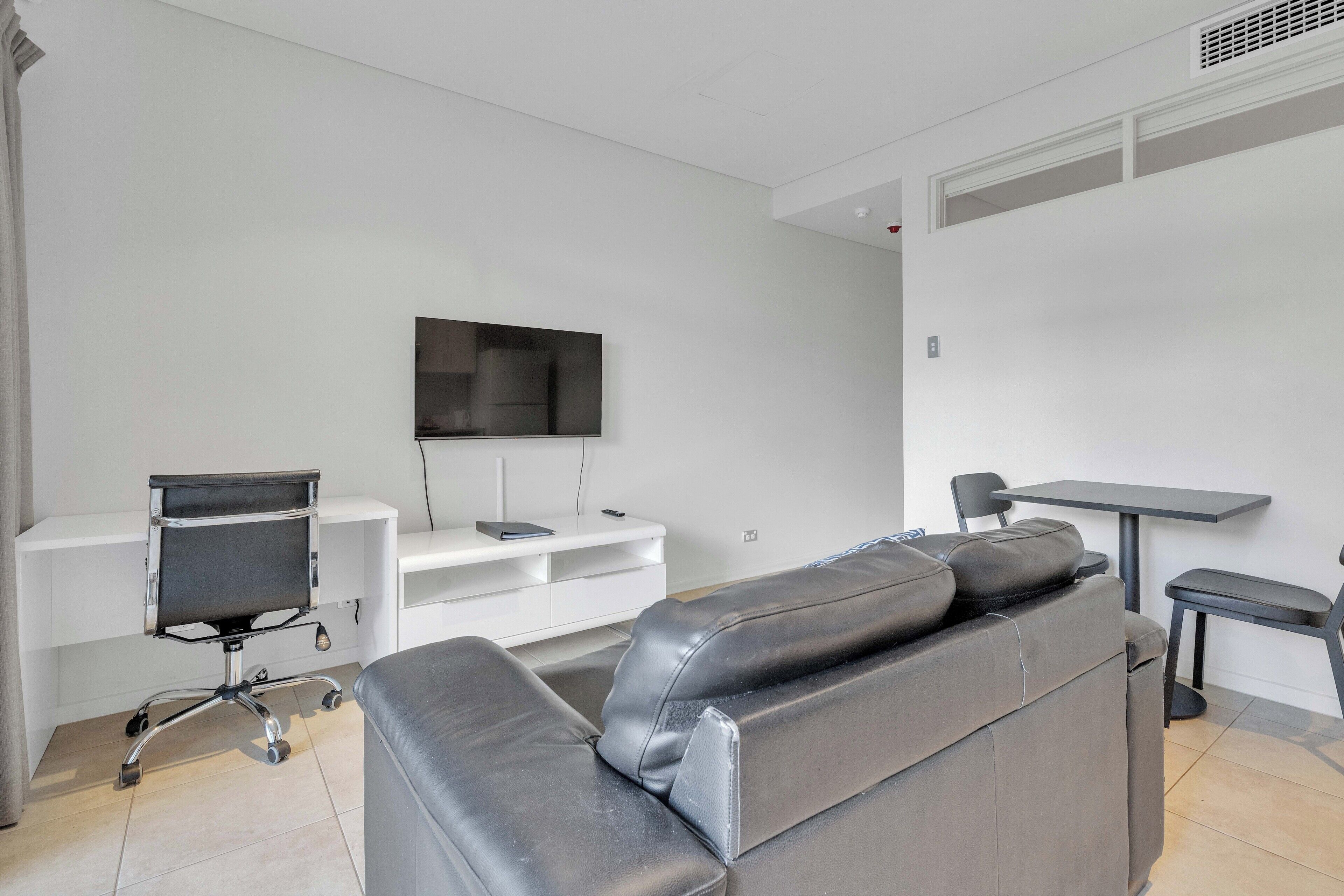 A modern living room with a leather sofa, a flat-screen TV mounted on the wall, a white entertainment unit, and a small table with a chair.