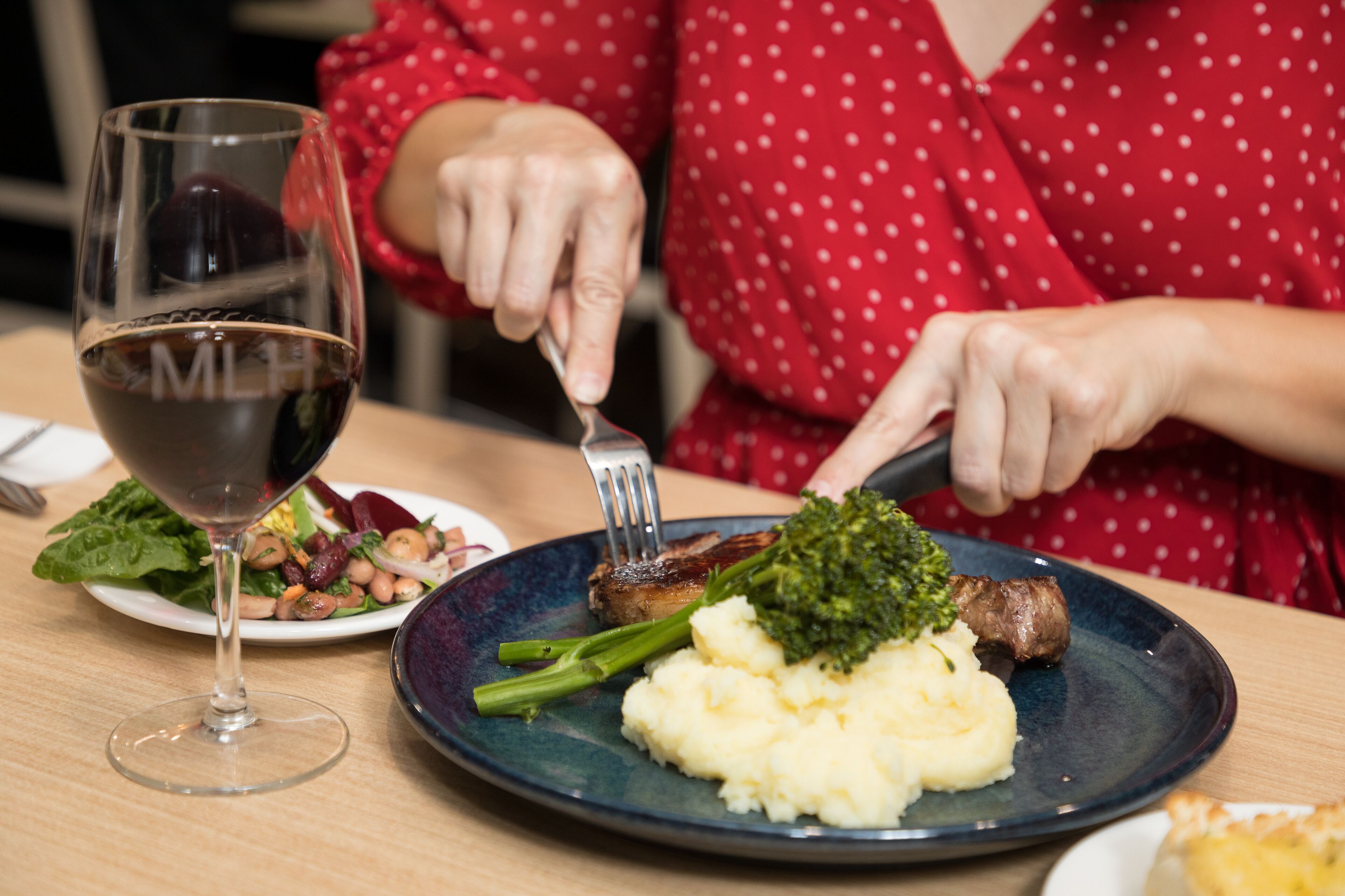 A dining table with a plate of food, a glass of red wine, and a person cutting meat.