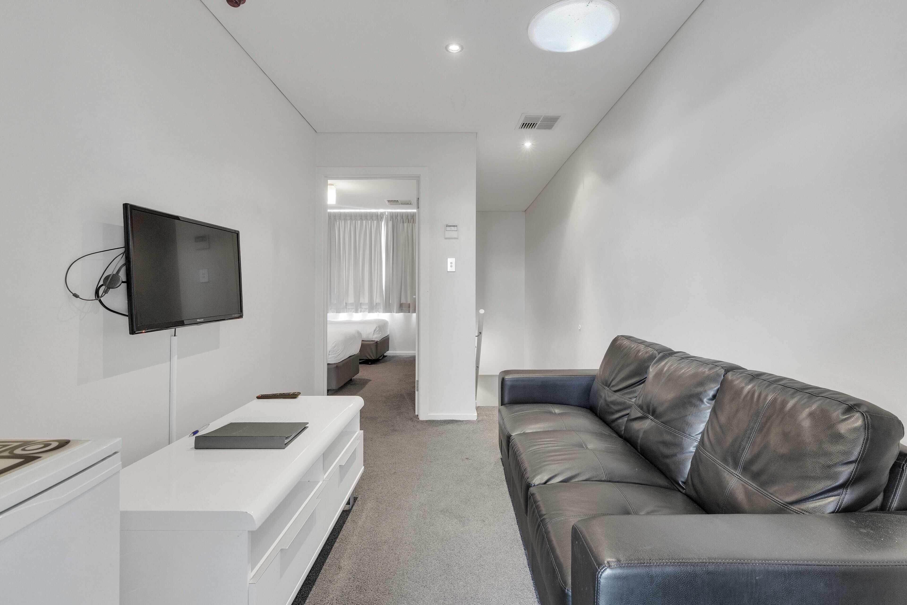 A modern hotel room with a flat-screen TV, a white desk, a leather sofa, and a separate bedroom with a bed and curtains.