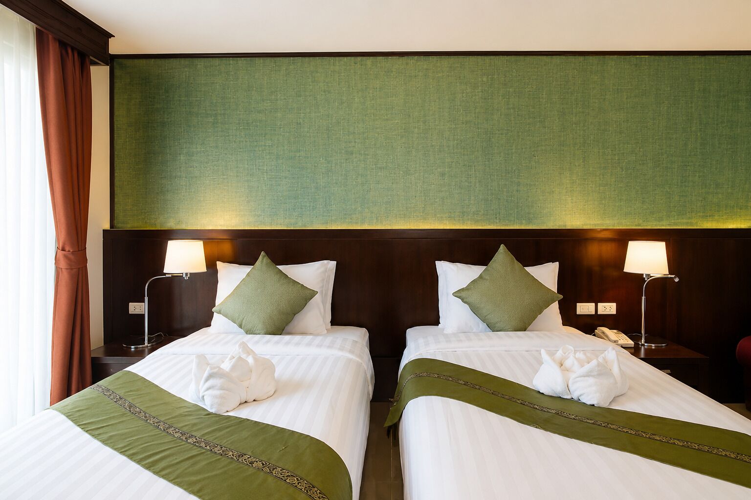 A hotel room with two beds, each with a green bedspread and pillows, a green headboard, bedside lamps, and a window with curtains.