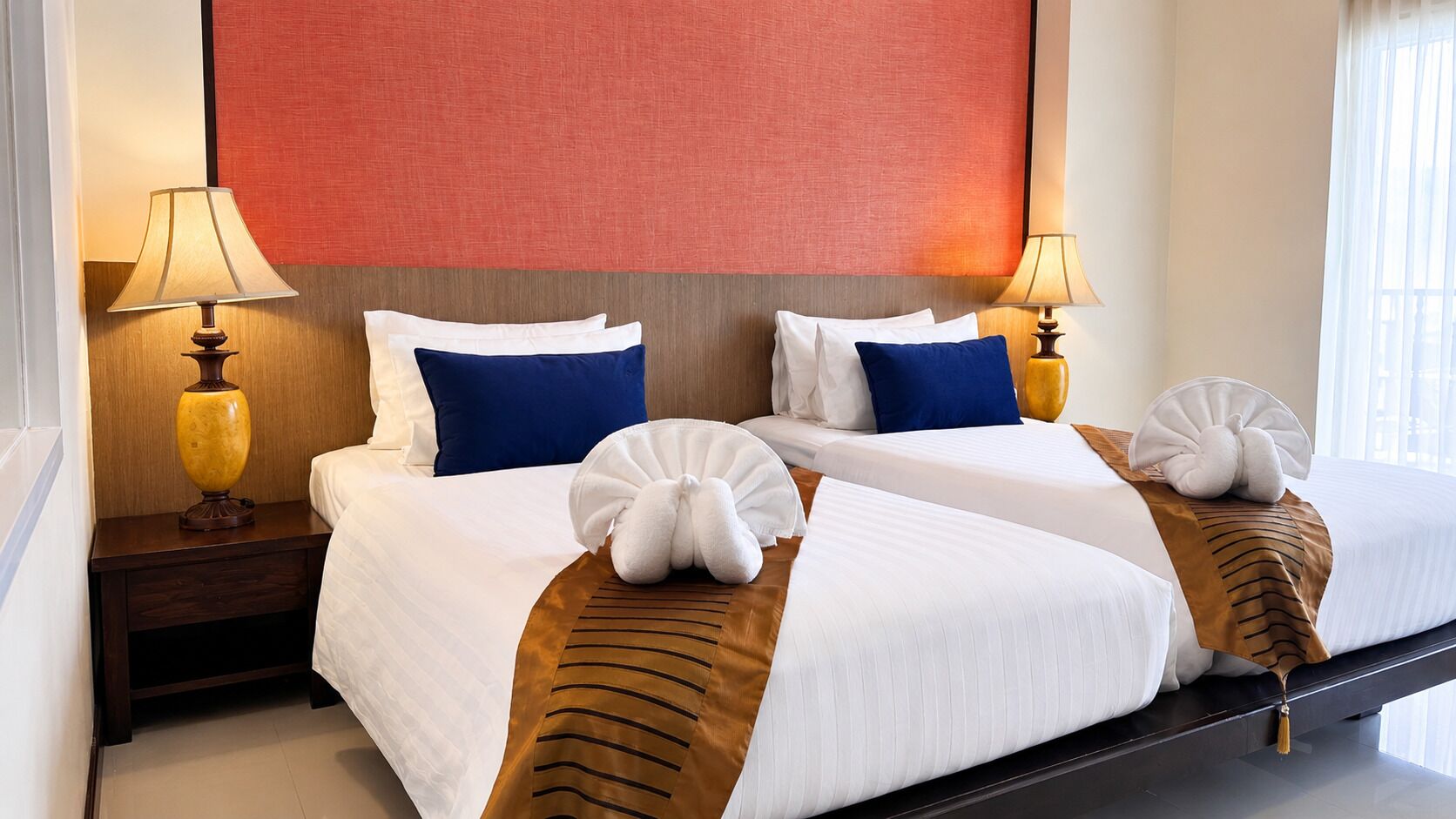 A hotel room with two beds, each with white linens, blue pillows, and a brown striped pillow. There are two bedside lamps, a red accent wall, and a window with curtains.
