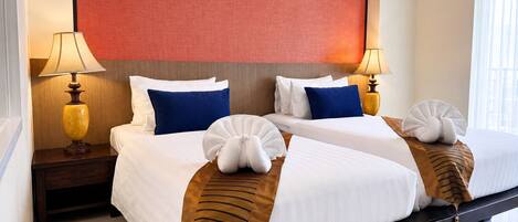 A hotel room with two beds, each with white linens, blue pillows, and a brown striped pillow. There are two bedside lamps, a red accent wall, and a window with curtains.