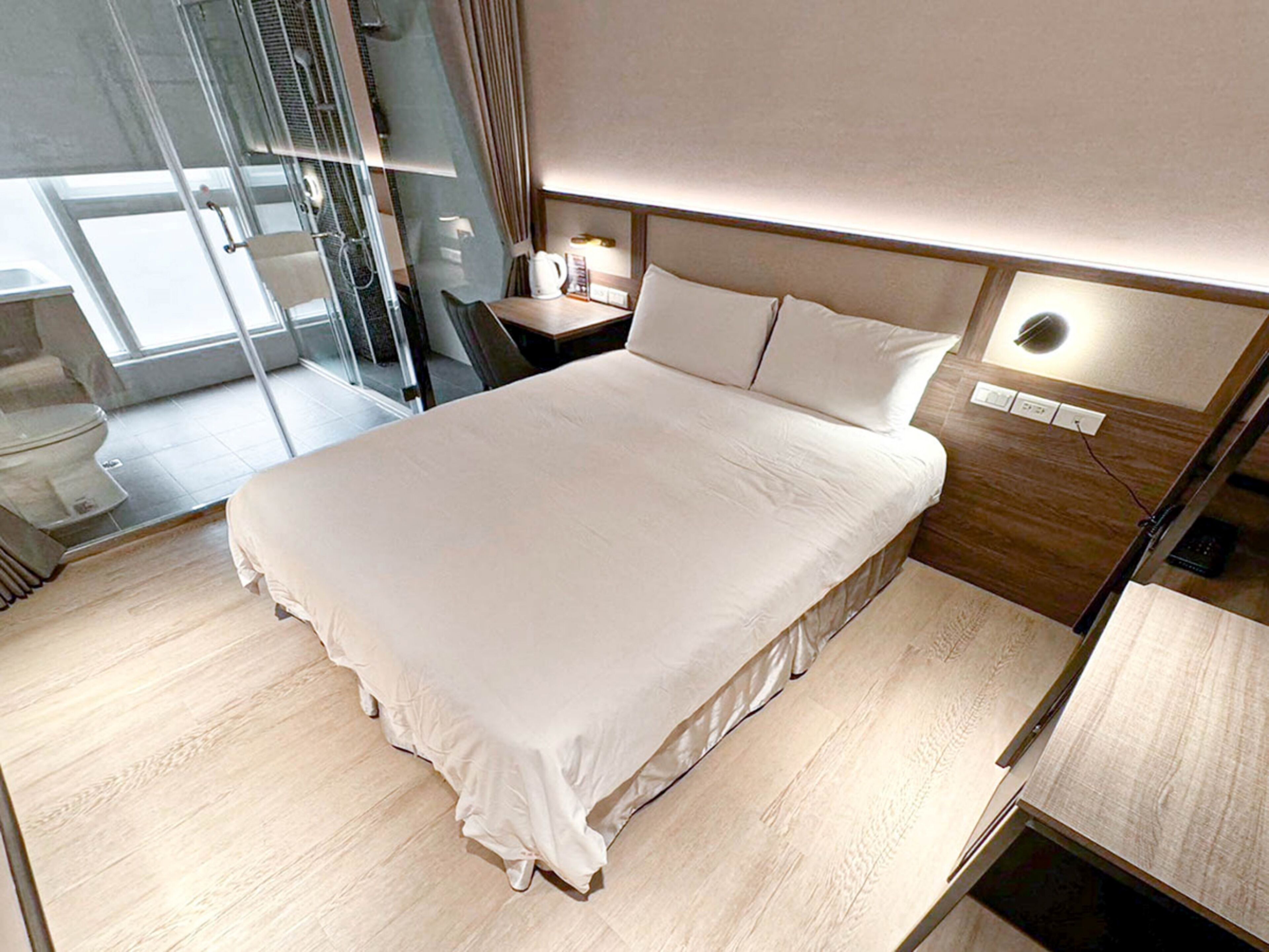 A modern hotel room with a large bed, a bedside table, a glass shower enclosure, and a view of the outside.