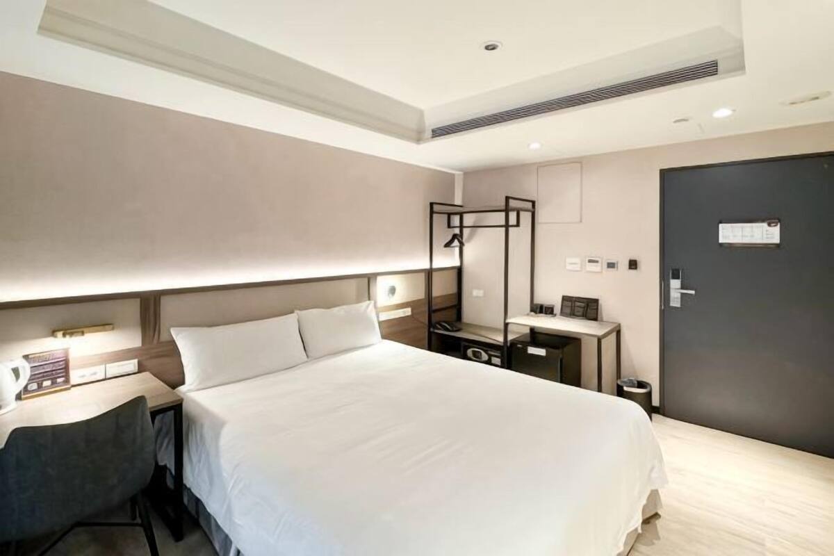 A hotel room with a large bed, bedside table, desk, chair, and a wardrobe.