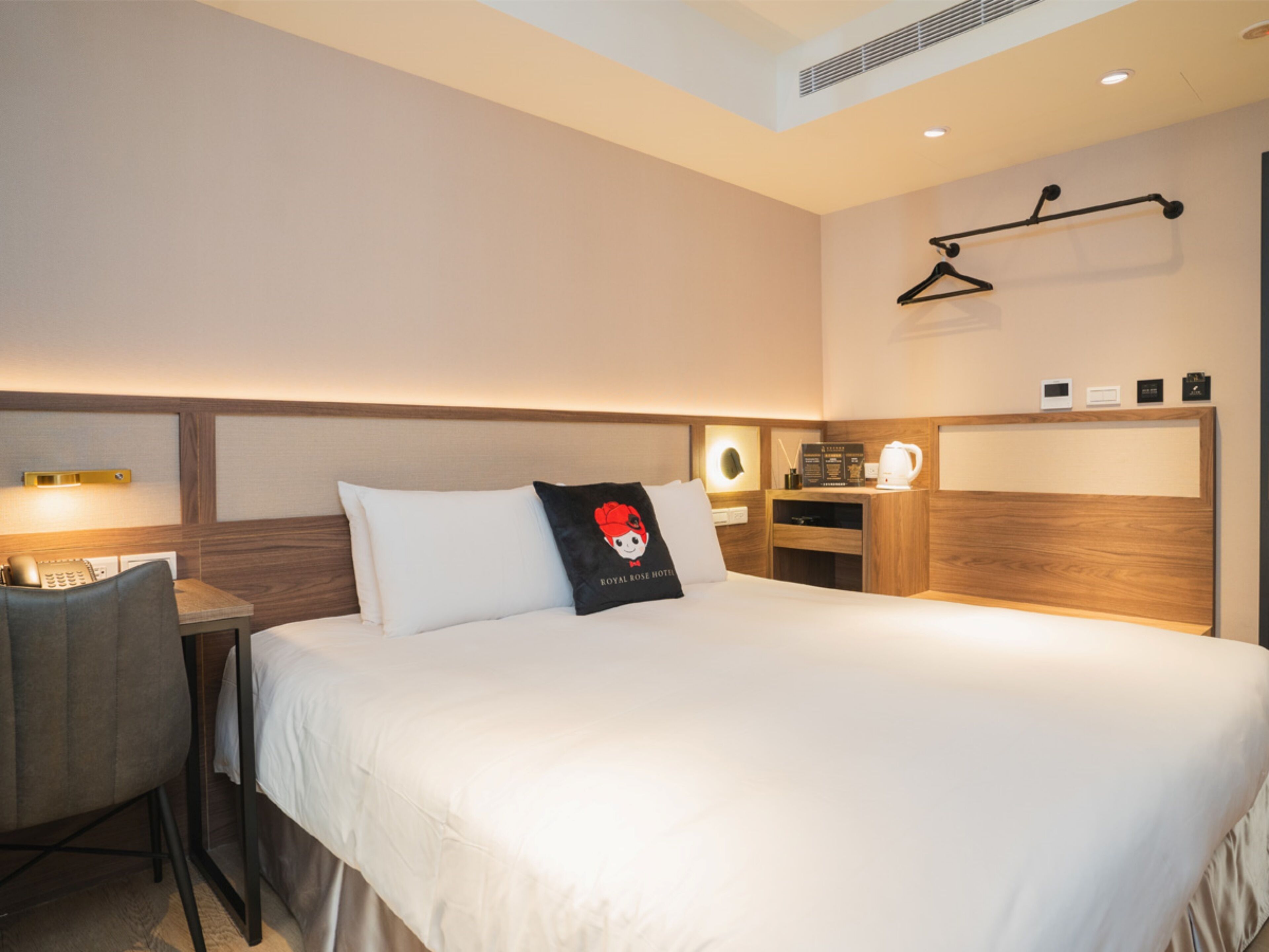 A hotel room with a bed, a desk with a chair, a bedside table, a wall-mounted heater, and a coat rack.