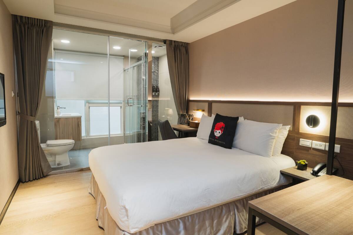 A hotel room with a large bed, a desk, a chair, a bedside table, a phone, and a window with curtains.