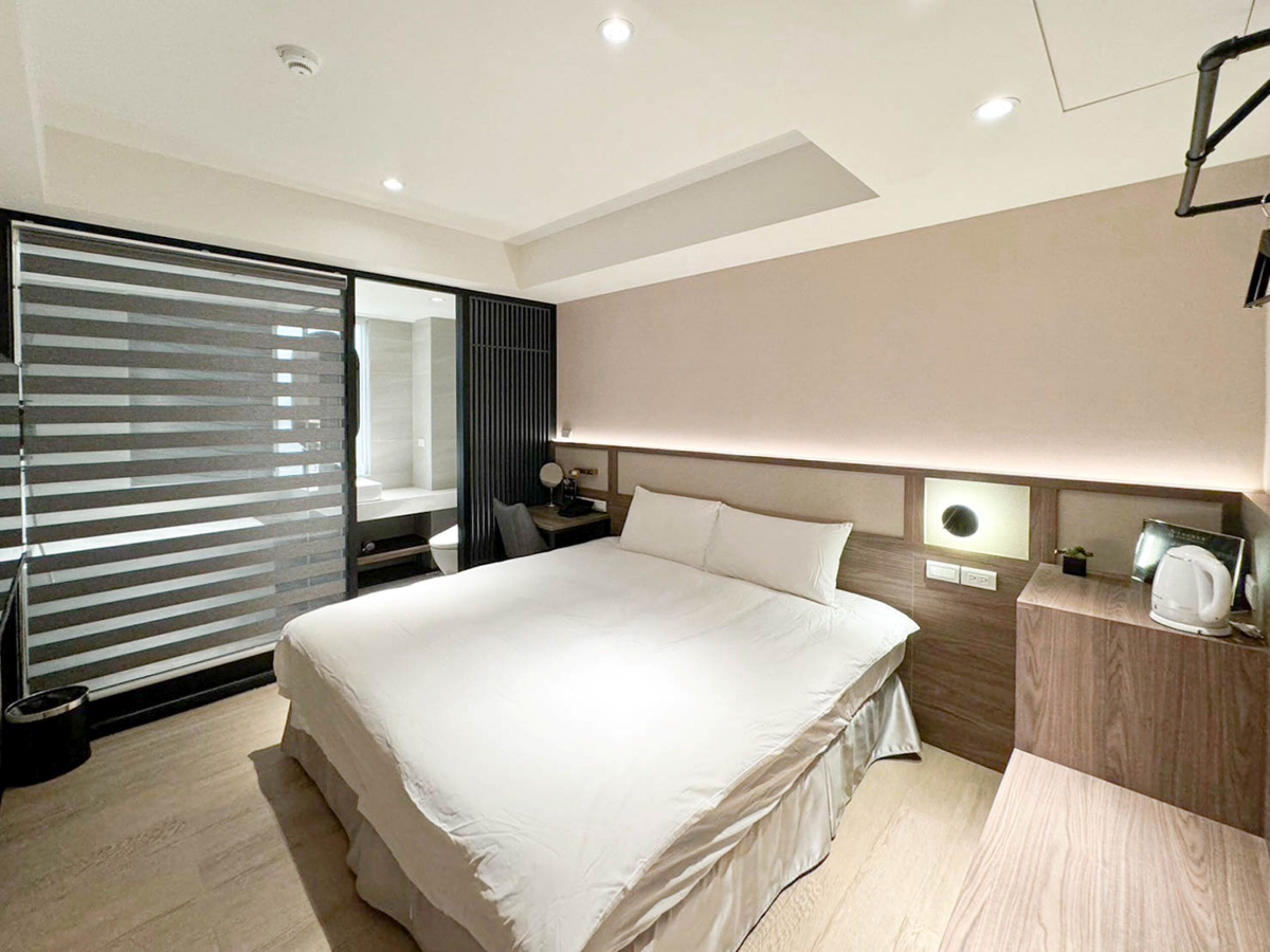 A modern hotel room with a large bed, a bedside table with a lamp, a small desk, and a built-in bench at the foot of the bed.