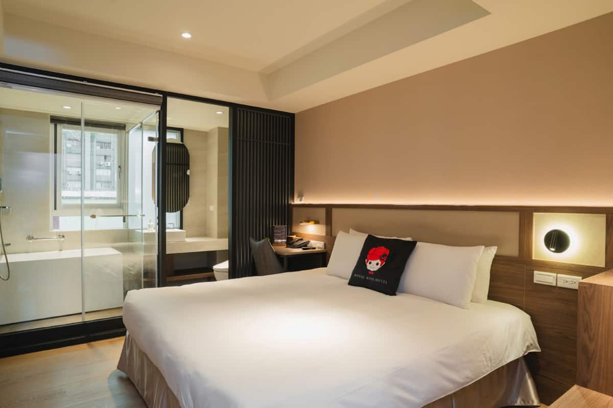A hotel room with a large bed, a desk, a chair, a nightstand, a lamp, and a glass shower enclosure.
