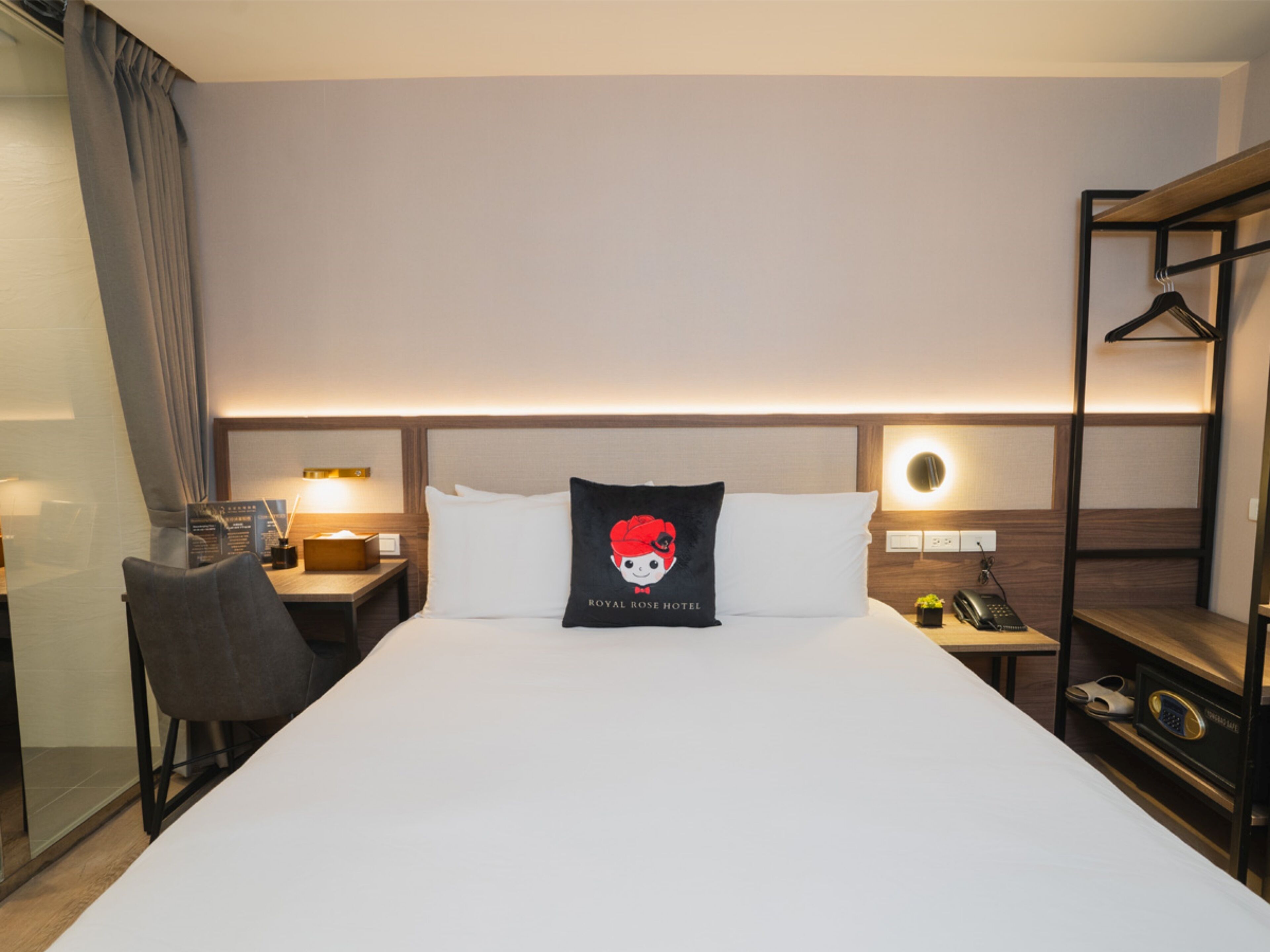 A hotel room with a large bed, a desk, a chair, a bedside table with a lamp, a telephone, and a coat rack.