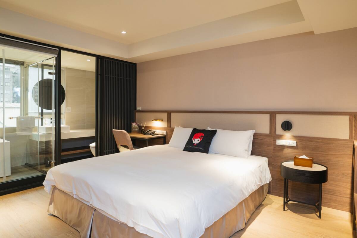 A hotel room with a large bed, a desk with a chair, a small table with a lamp, and a glass partition.