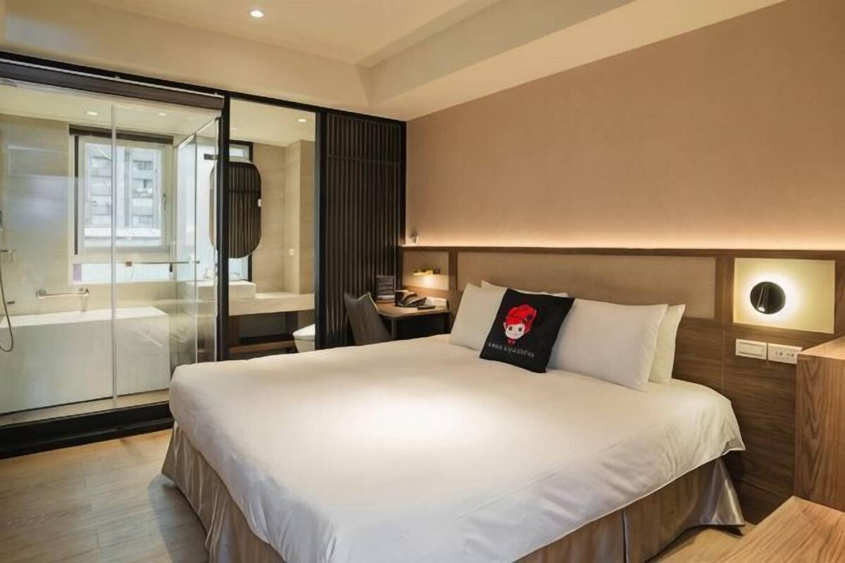 A hotel room with a large bed, a glass shower enclosure, a bedside table with a lamp, and a wall-mounted clock.