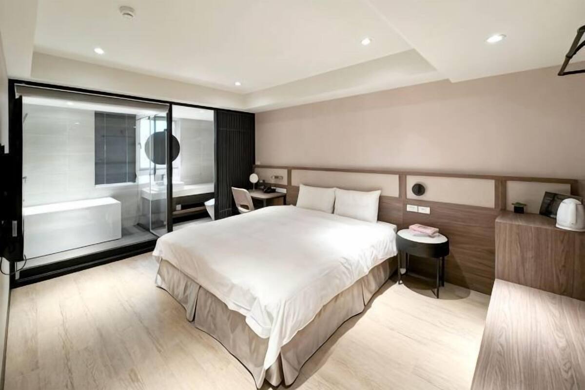 A modern hotel room with a large bed, a desk, a chair, a small table, a nightstand, and a sliding glass door leading to a balcony.
