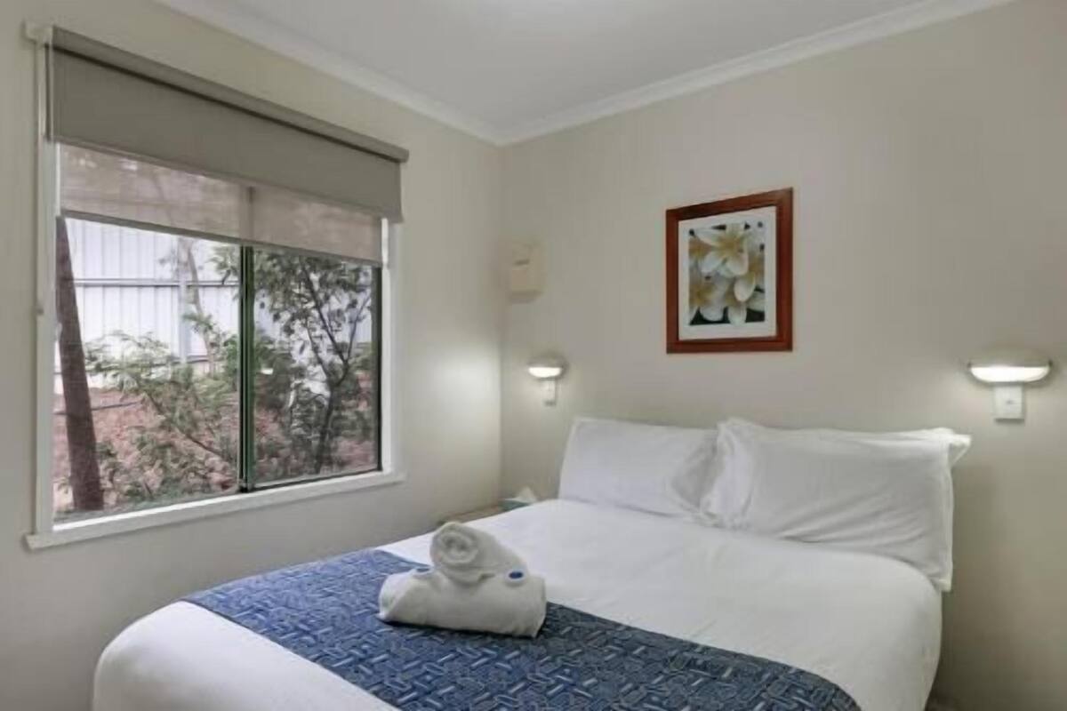 A hotel room with a bed, a window with a view of trees, a framed picture on the wall, and two wall-mounted lights.