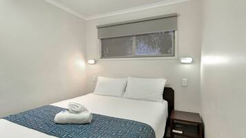 A neatly made bed with a patterned bedspread and a rolled towel, a small bedside table, and a window with a blind.
