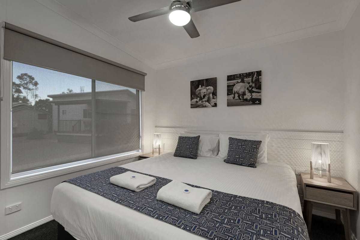 A hotel room with a large bed, two wall-mounted nightstands, a ceiling fan, and a window with a view of buildings.