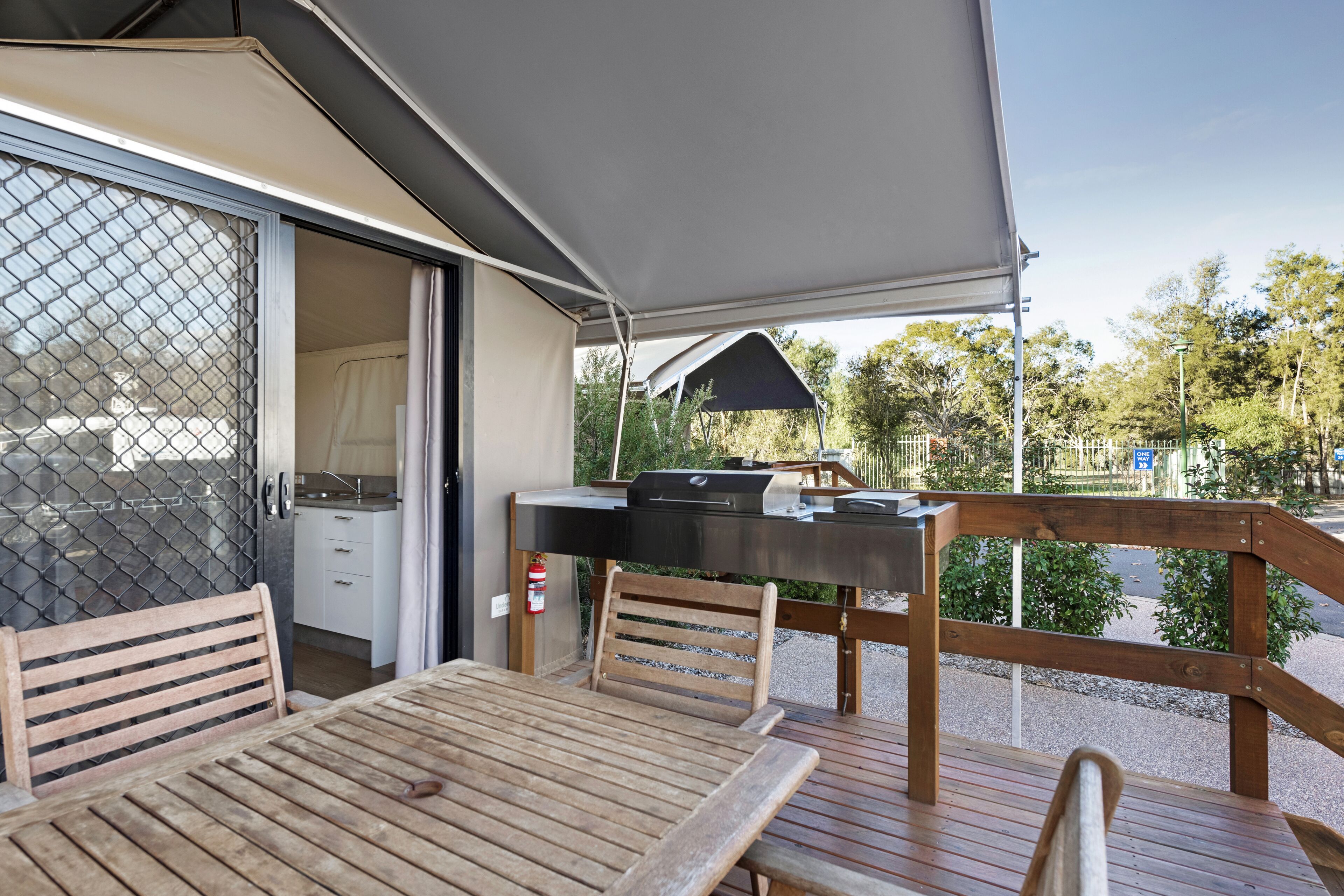 Una terrazza esterna con tavolo e sedie in legno, un barbecue e una zona riparata con vista sulla strada e sugli alberi.