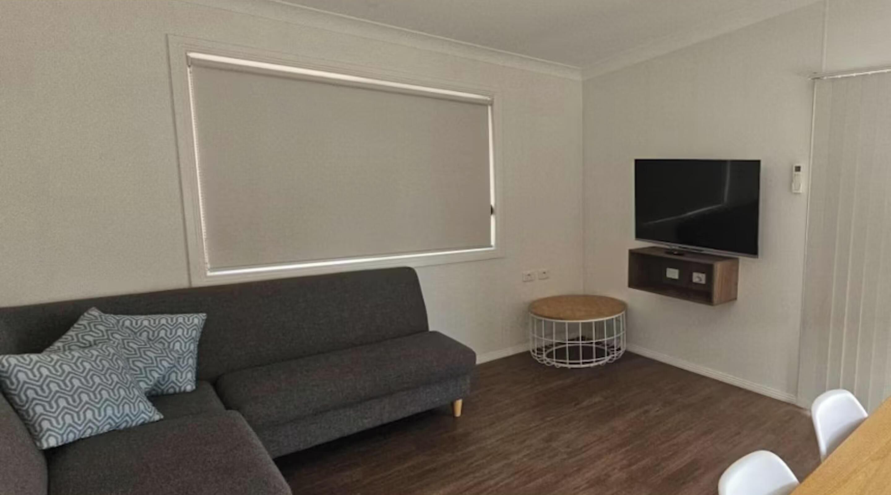 Ein modernes Wohnzimmer mit grauem Sofa, einer runden Schlafgelegenheit für Haustiere, einem flachbildfernseher an der Wand und einer Essecke mit weißen Stühlen.
