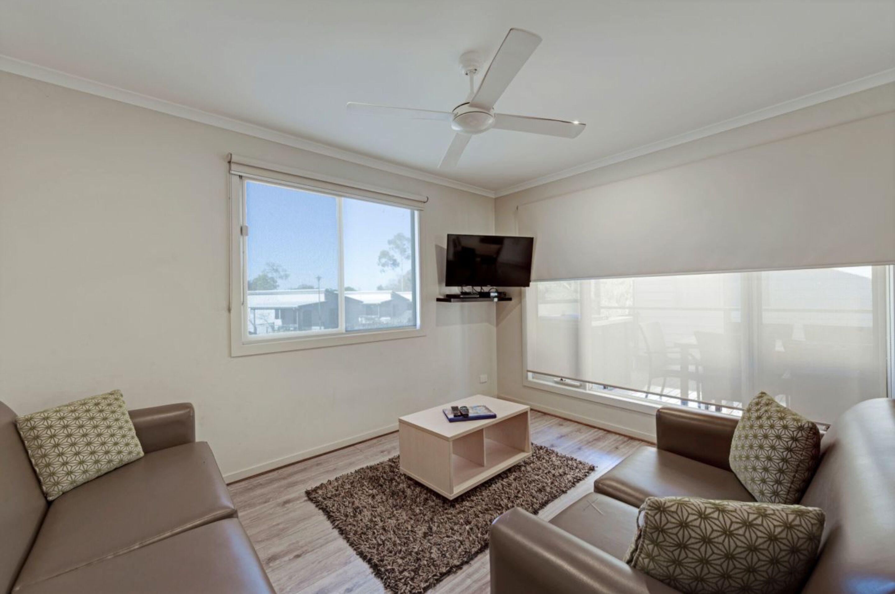 Una sala de estar moderna con ventilador de techo, televisor de pantalla plana montado en la pared y un ventanal con vistas al exterior.
