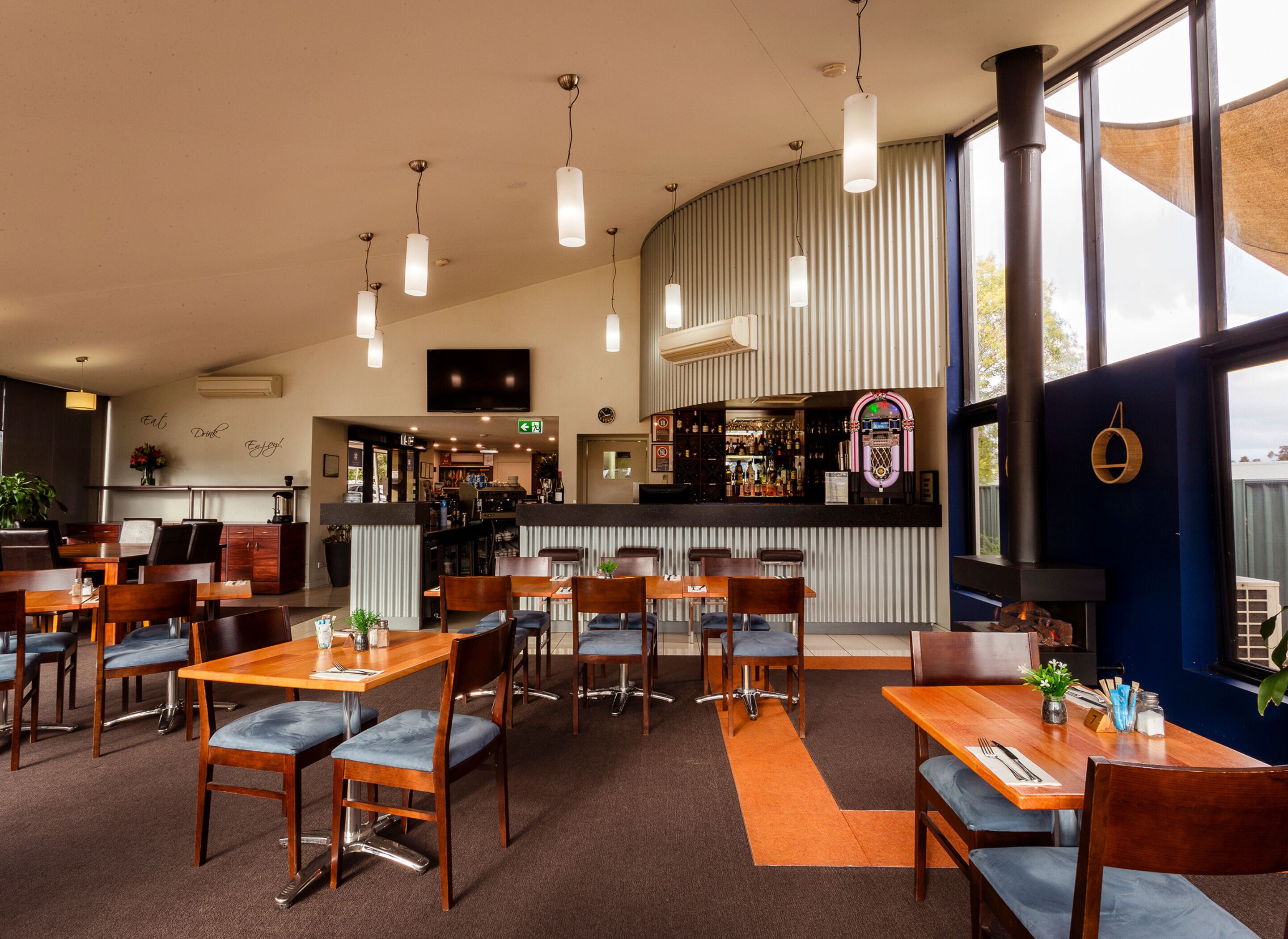 Un restaurante con varias mesas y sillas, una zona de bar y un amplio ventanal que proporciona luz natural.