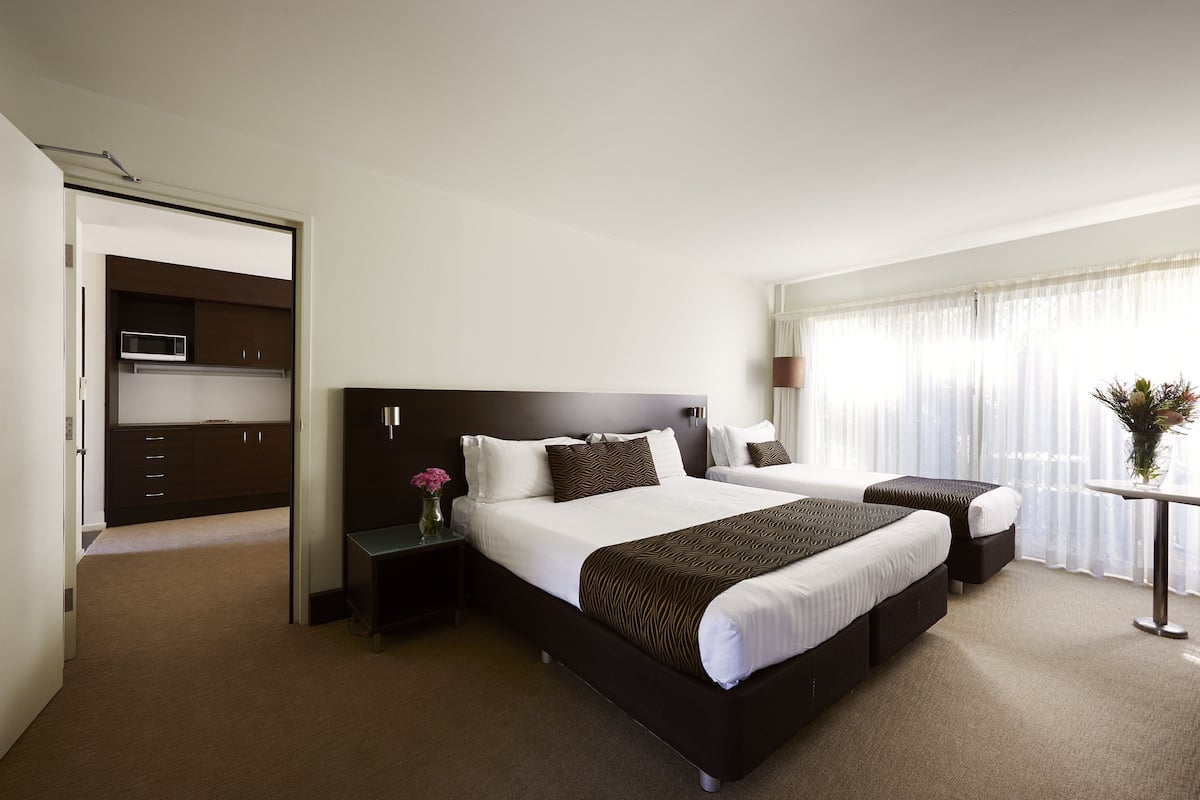 A hotel room with a large bed, a smaller bed, a desk, a chair, a nightstand, a microwave, and a window with curtains.