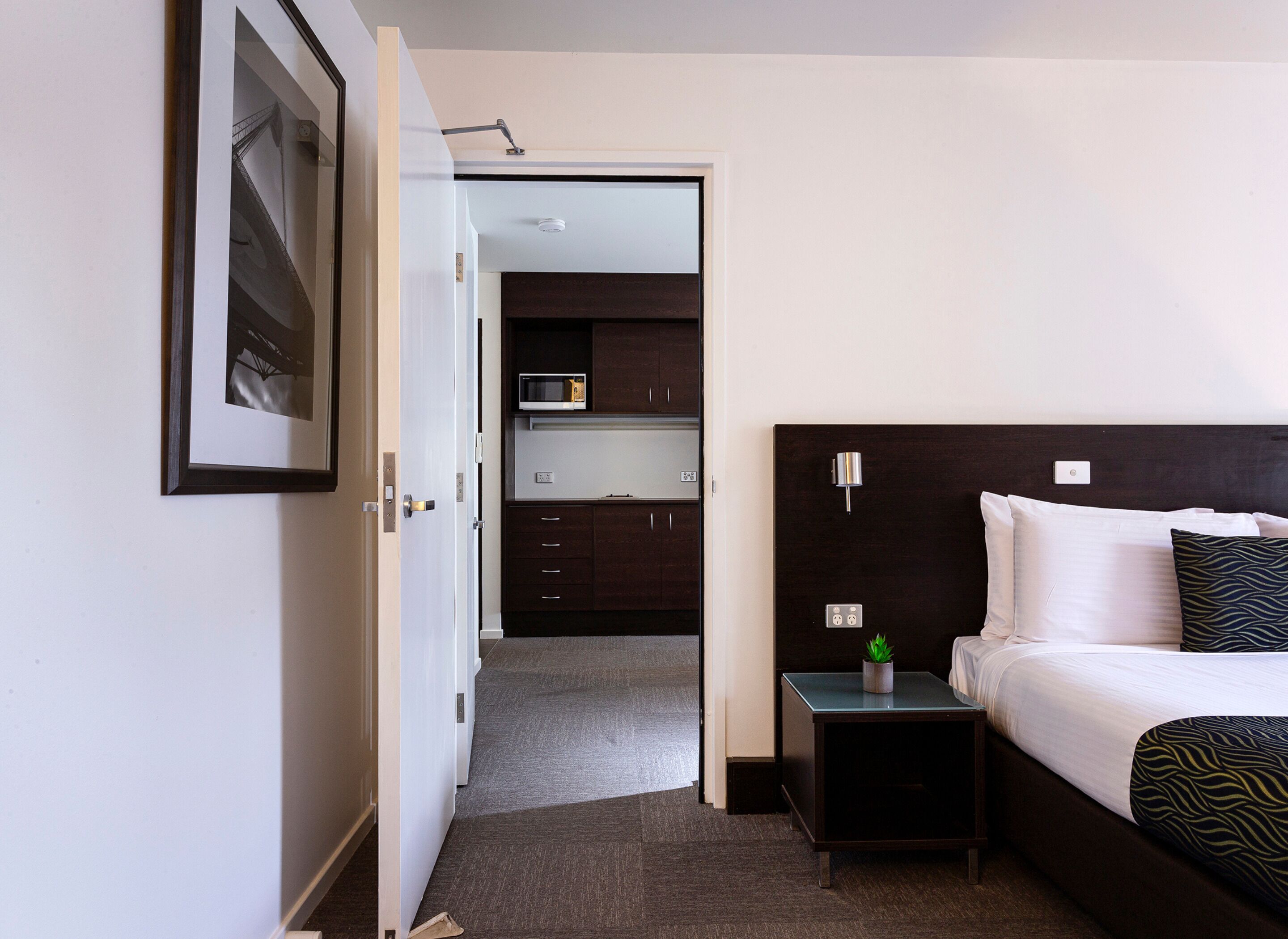 A hotel room with a bed, bedside table, and a view into the kitchen area.