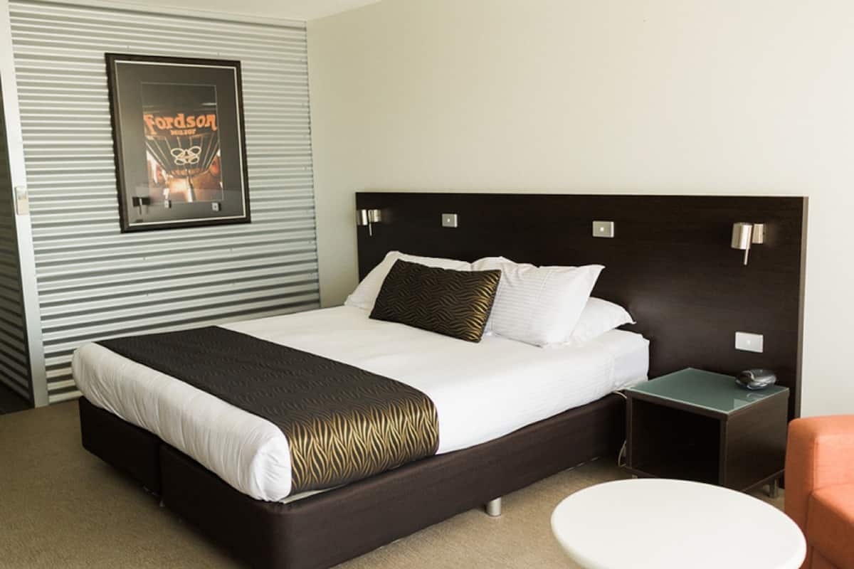 A hotel room with a large bed, a bedside table, a chair, and a poster on the wall.