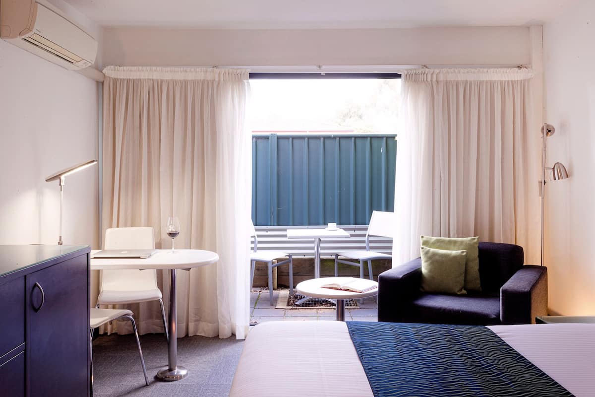 A hotel room with a bed, a desk with a chair, a small table with a chair, a sofa, and a window with curtains.