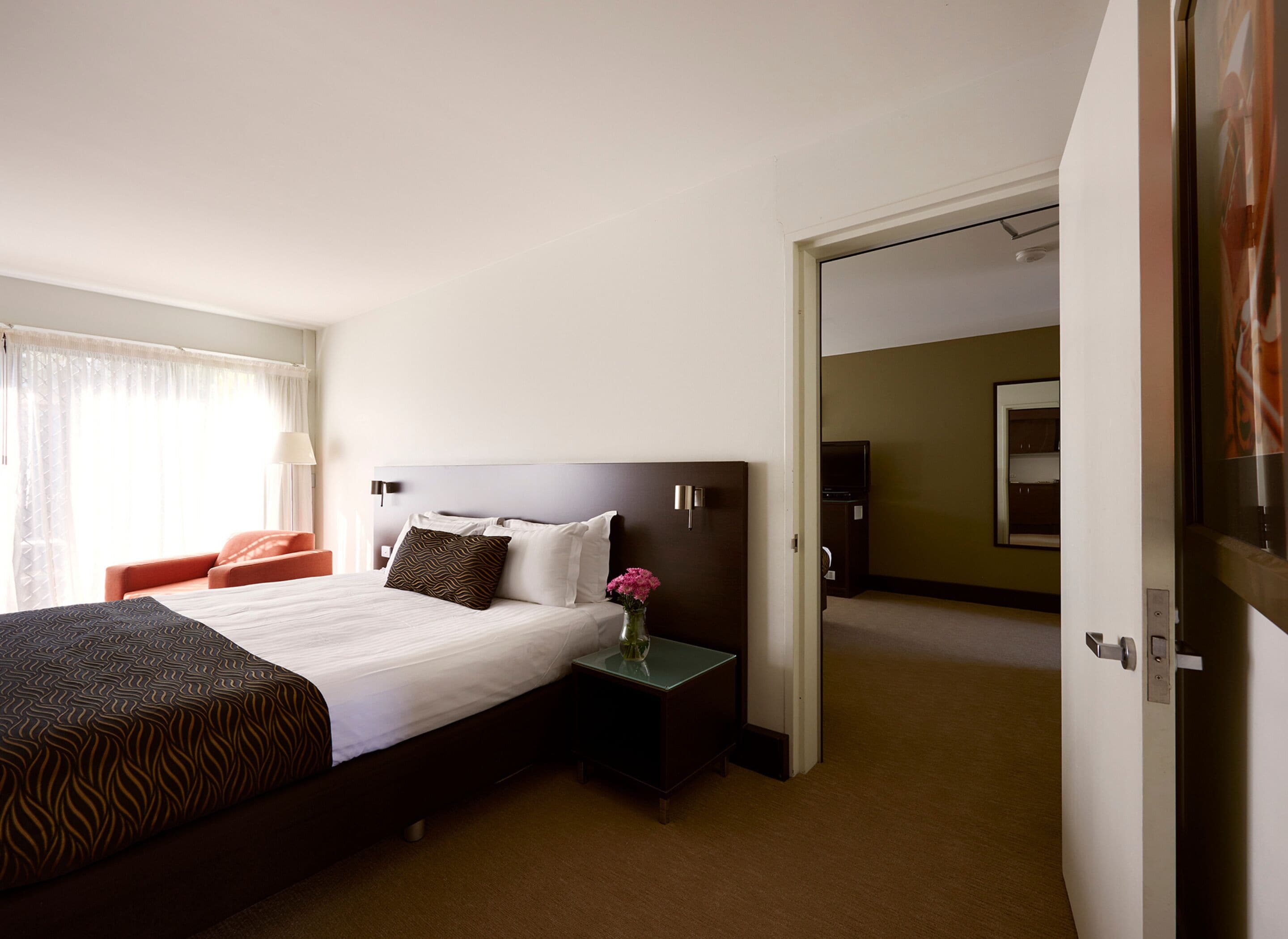 A hotel room with a large bed, a nightstand with a vase of flowers, a desk, and a view of the hallway.