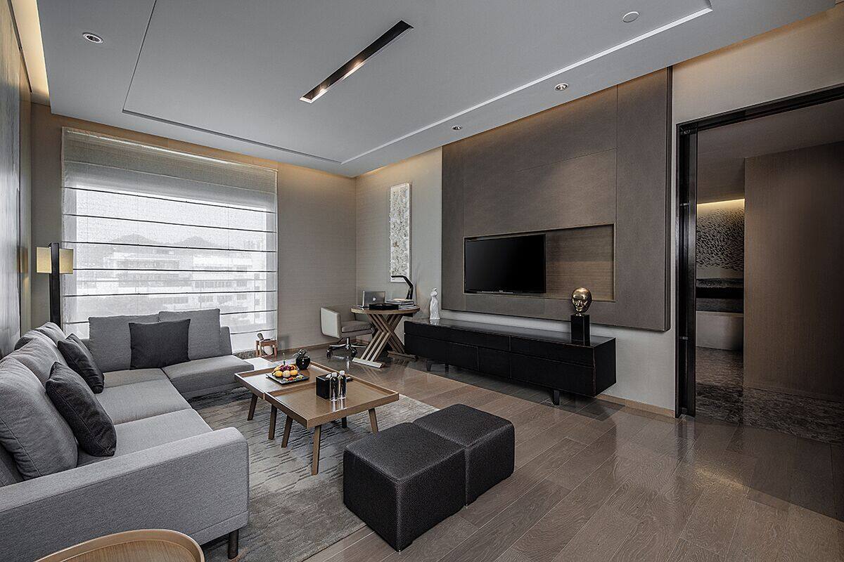 A modern living room with a sectional sofa, a coffee table, a flat-screen TV mounted on the wall, and a small side table with a lamp.