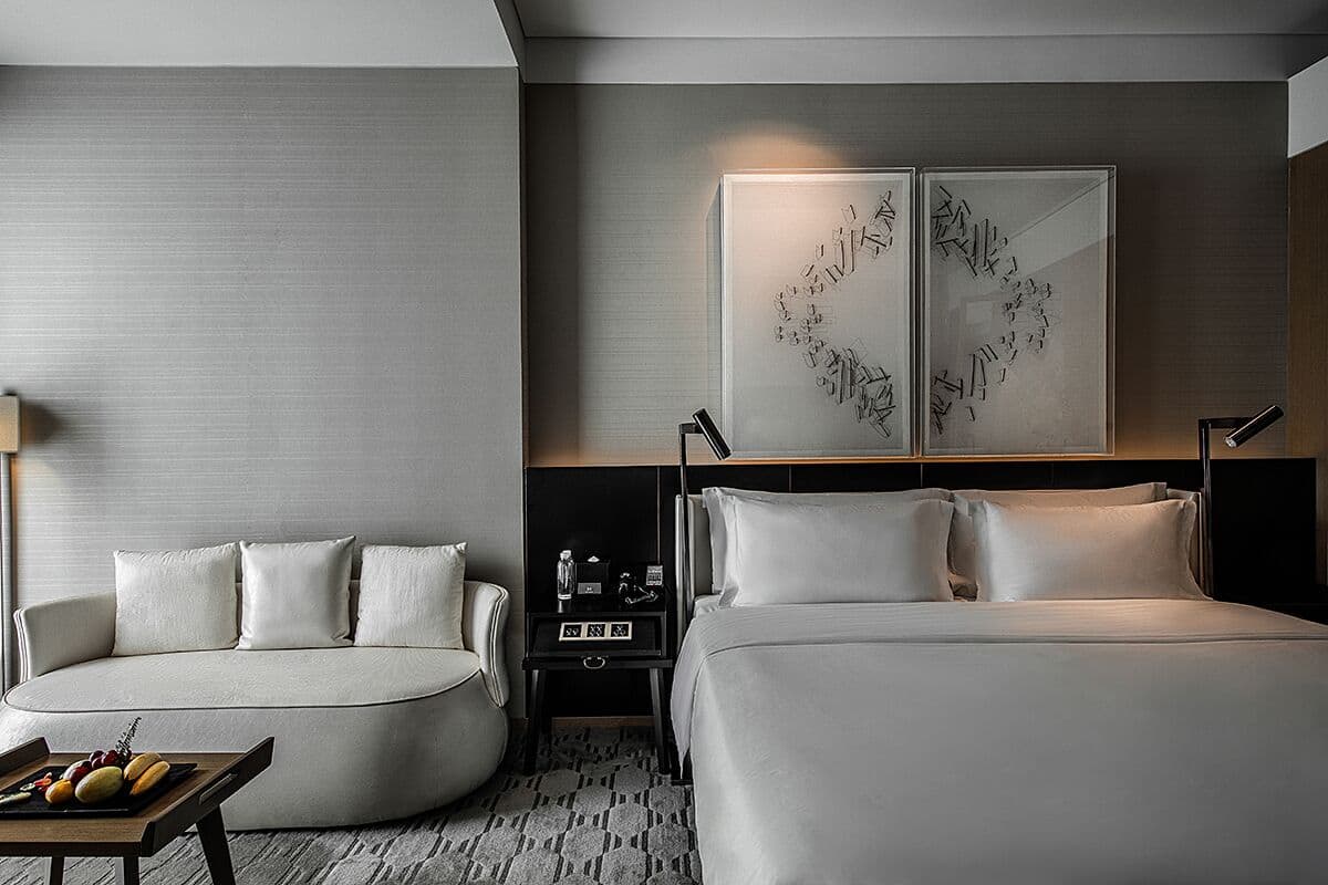 A modern hotel room with a curved sofa, a bed, a nightstand with a lamp, and artwork on the wall.