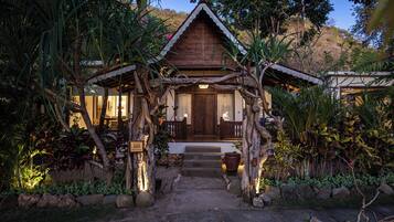 Une maison en bois avec un porche, entourée de plantes tropicales et un chemin de pierres menant à l’entrée.