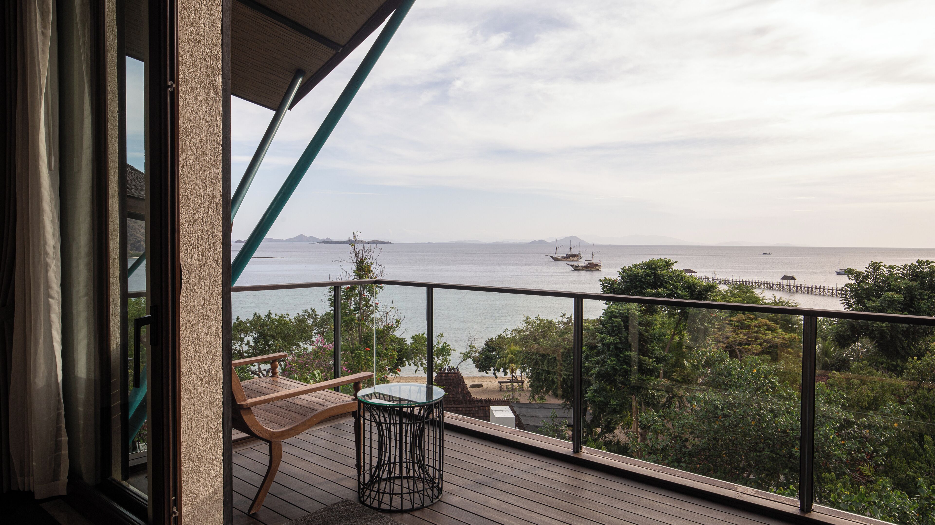 A balcony with a view of the sea, wooden furniture, and a glass table.